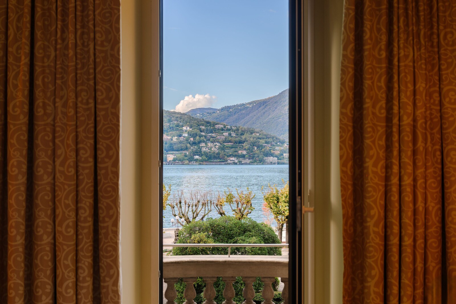 A view from a balcony with patterned curtains, overlooking a lake and a hillside with buildings. - Palace Hotel (Como)